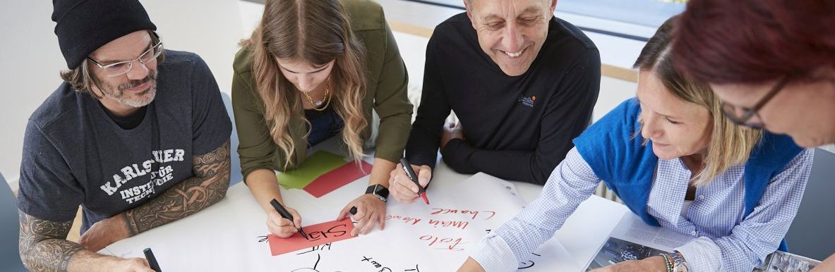 Gruppe von Personen arbeitet gemeinsam an einem großen Whiteboard-Tisch.