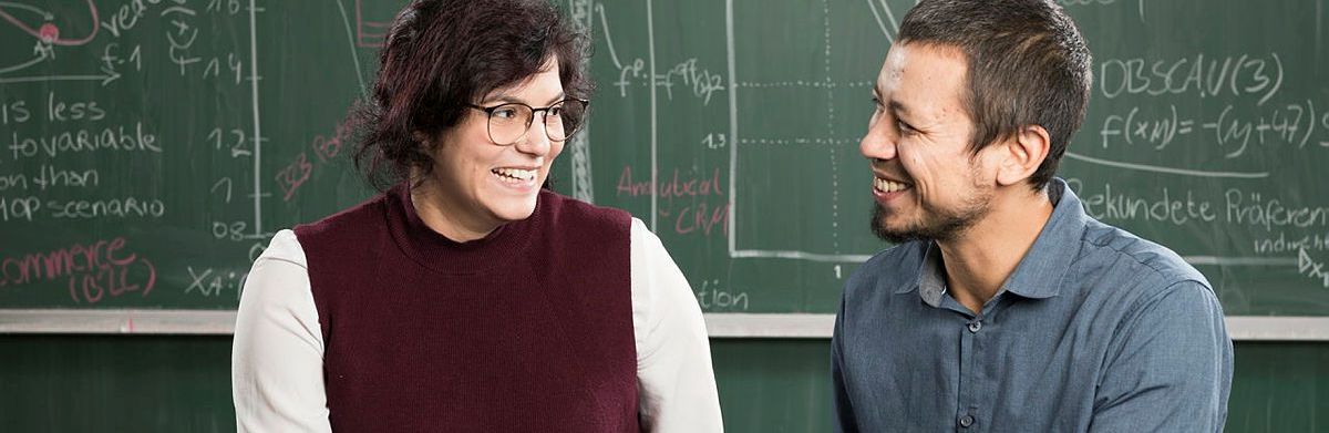 KIT Zwei Personen besprechen sich in einem Seminarraum vor einer Tafel mit mathematischen Formeln,
